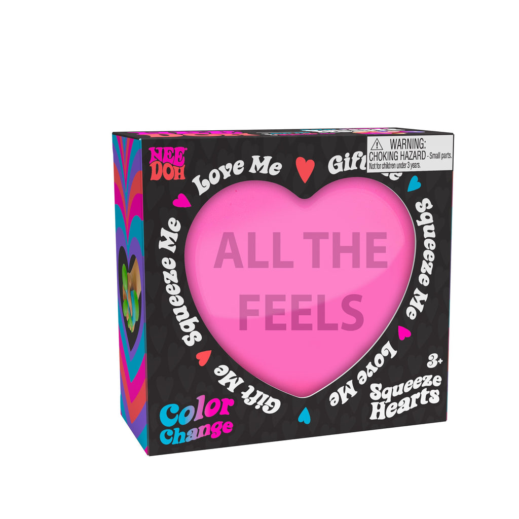 A boxed Nee Doh Color Change Squeeze Heart E1 toy. The packaging is predominantly black with colorful accents, featuring phrases like "ALL THE FEELS," "Squeeze Me," "Love Me," and "Gift Me." The box also includes a choking hazard warning for small parts.